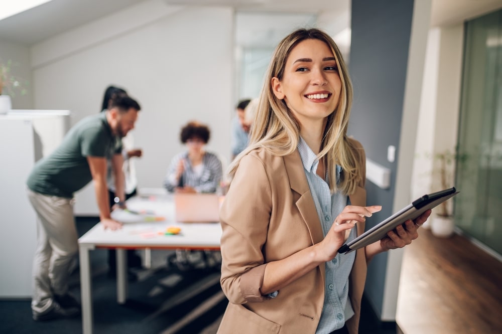 Femme d'affaires avec tablette dans un bureau moderne Jeune femme souriante en blazer utilisant une tablette dans un bureau moderne avec ses collègues en arrière-plan.