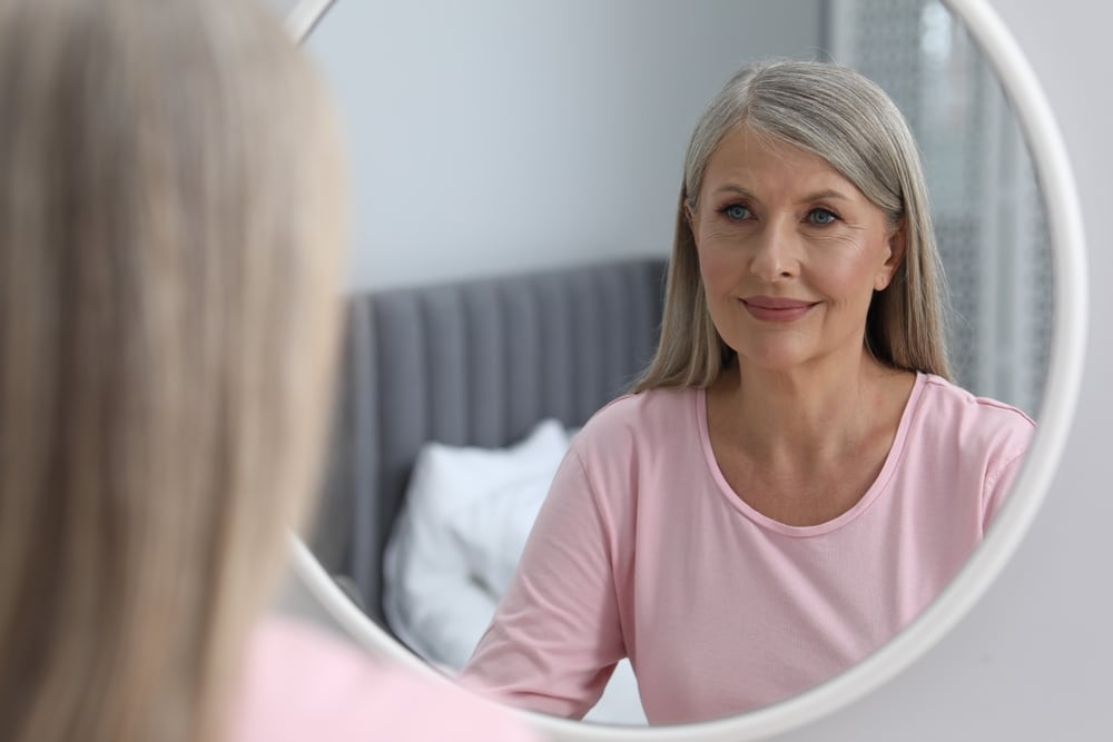 Femme mûre aux cheveux gris souriant à son reflet dans un miroir rond, vêtue d'un t-shirt rose.